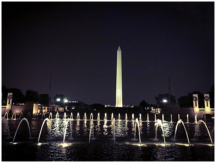 The WWII Memorial fountain lit up at night with a lit Washington Monument in the background.