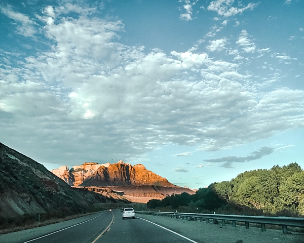 A white car drives along a road heading towards a large rock formation near Zion National Park